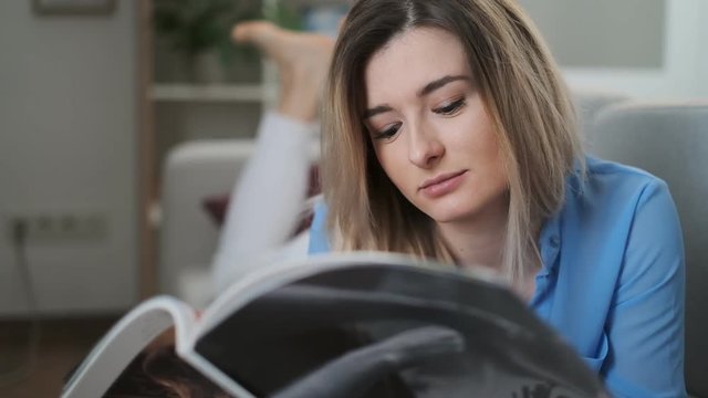 Young charming girl flipping a fashion magazine pages while lying on the couch in the cozy living room. Woman relaxing at home, she is sitting on the sofa and reading a catalog. Close up