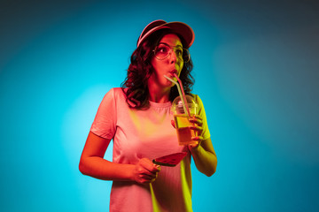 Chilling young woman in a cap holding sweets and drink over trendy blue neon studio background. Beautiful female portrait. Concept of human emotions, facial expression, summer holidays or weekend.