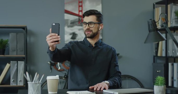 Good looking smiled Caucasian man in glasses and business style talking while having videochat on the smartphone in the cozy ofice room.