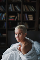 Obraz premium Pensive beautiful young woman with blond short hair sitting on bed on white linen. The impression of tenderness and innocence. Sunny and calm morning. In the background is a dark bookcase.