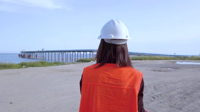 Giovane ingegnere donna con gilet arancione e caschetto bianco va a lavorare al pontile sulla spiaggia di fronte all'oceano.