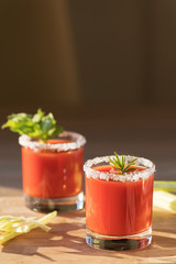 Beautiful tomato juice in glasses with celery, salt and rosemary on a wooden cutting board on a bright sunny day