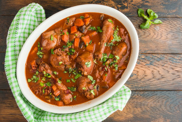 Coq au Vin, traditional French recipe of chicken braised in red wine with carrot and mushrooms. White casserole on wooden table, top view