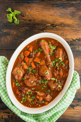 Coq au Vin, traditional French recipe of chicken braised in red wine with carrot and mushrooms. White casserole on wooden table, top view