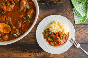 Coq au Vin, traditional French recipe of chicken braised in red wine with carrot and mushrooms. Served with mashed potatoes. White casserole and plate on wooden table, top view