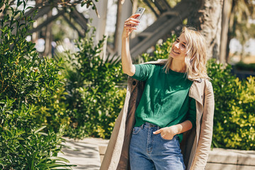 Beautiful young woman smiling while holding smartphone strolling outdoor in the city. Happy female taking selfie on mobile phone. Pretty girl making self portrait with fun face on cellphone. Travel