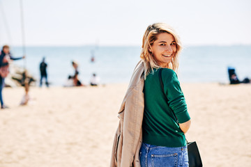 Portrait of beautiful blonde woman smiling and enjoying the warm weather on the beach. Pretty female walking on the beach against calm sea, blue sky background with copy space for your content. Travel