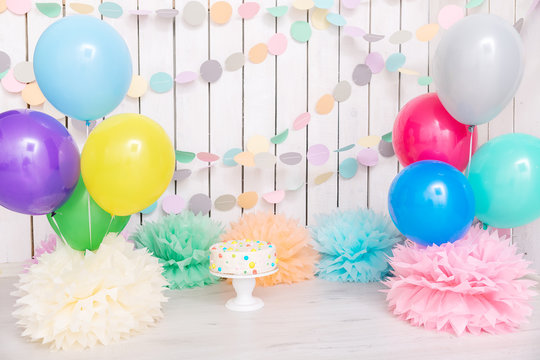 Photo zone with paper garlands, balloons, paper balls, pom poms, confetti and cream cake. Birthday cake. Smash cake. One year. Pink, white, blue, green, yellow colors. Rainbow