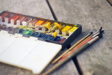 Naklejka premium On a gray wooden table are vintage watercolor paint in a box with cuvettes, along with which are yellow flowers coltsfoot and a few brushes for drawing.