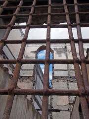 view through rusty iron bars on a window of a ruined collapsing building with crumbling walls and open to the sky