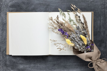 Opened blank sketchbook mockup with white pages and spring bouquet of dried flowers and herbs wrapped in craft paper lying on a dark chalkboard.