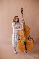 Beautiful young woman musician sitting on a vintage double bass on a beige background in a studio © polinaloves