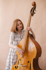 Beautiful young woman musician sitting on a vintage double bass on a beige background in a studio © polinaloves