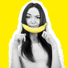 Young woman holding banana like smile near her face over yellow background.