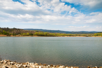 A bright day at the spring lake with light clouds against the background of the forest and beautiful mountains