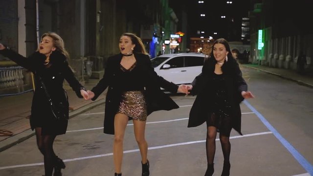 Girls night out. Special event. Ladies in sparkling dresses holding hands, running to party. Street city lights background.