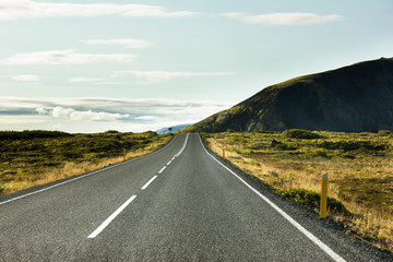 Leaving into the distance highway, road with grass and moss on the sides, blue sky and clouds and green mountain, hill in the distance. The great nature of Iceland. Road trip around Iceland island.