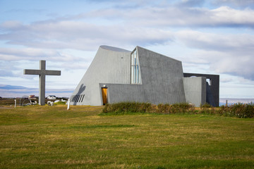 Unusual gray church in the middle of the meadow and green grass in the north of Iceland. Modern Scandinavian Icelandic church architecture. Summer trip around Iceland. Cross standing near the church.