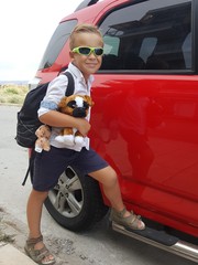 A boy-backpacker in sunglasses is ready for adventures, standing at a red car
