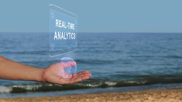 Male hands on the beach hold a conceptual hologram with the text Real-time analytics. A man with a holographic technology on the background of the sea