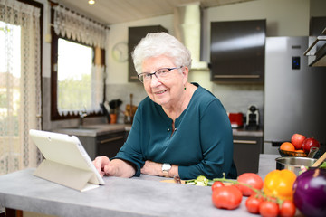 happy senior active woman cooking at home in modern kitchen and looking for recipe on a internet computer tablet