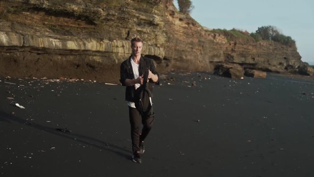 Man with backpack walks along black sand beach, does acrobatic trick. Slow motion, steadicam shot