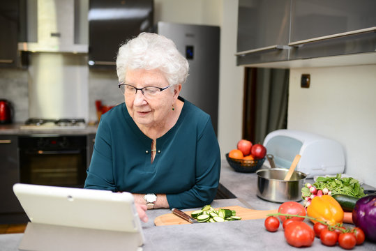 Happy Senior Active Woman Cooking At Home In Modern Kitchen And Looking For Recipe On A Internet Computer Tablet