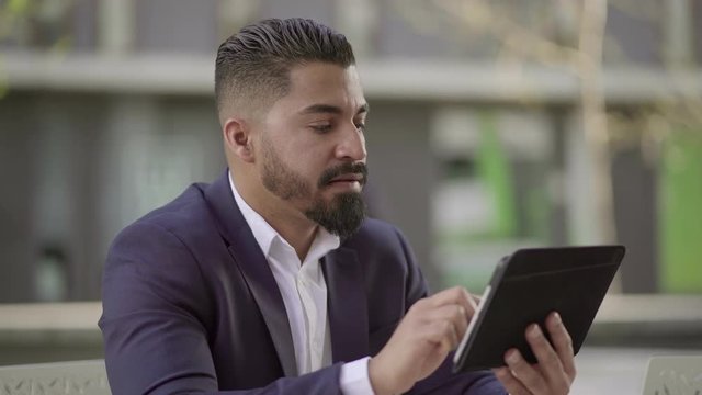 Serious handsome businessman using tablet pc outdoor. Focused man in suit jacket and white shirt sitting at outdoor cafe and working with digital tablet. Technology concept