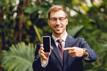 smiling businessman in suit, tie and glasses pointing with finger at smartphone blank screen in greenhouse