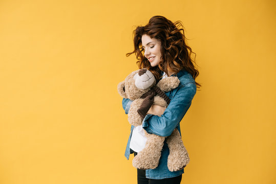 cheerful curly young woman holding teddy bear on orange