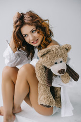 happy curly young woman holding teddy bear while touching hair on white