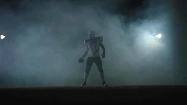 American football sportsman player in football helmet standing on the field on black background with spotlight in a cloud of smoke with the ball in hands
