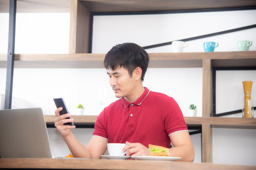 The business man with casual  red t-shirt messaging on mobile phone, drinking coffee and eating sandwich, Young man working on labtop or computer  in the kitchen room, loft style