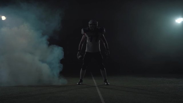 American football sportsman player in football helmet standing on the field on black background in a cloud of smoke with the ball in hands. The man raising ball makes invoking gestures