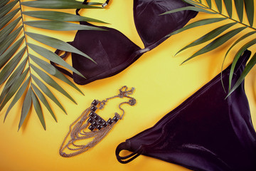 Bikini black velvet swimsuit, necklace and tropical palm leaves on yellow background. Overhead view of woman's swimwear.