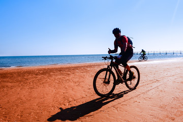 Fototapeta premium Man cycling on sport bike showing thumb up on ocean background - Silhouette of people riding bicycle on the beach shore with morning backlight - Lifestyle concept of healthy holiday activity - image