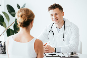 selective focus of cheerful doctor in white coat sitting with clenched hands and looking at patient