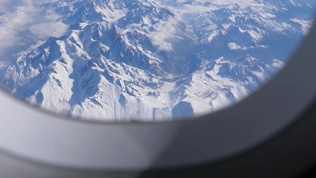 Looking through window at snow covered European Alps mountain range from flying aircraft showing part of porthole frame and clear view after camera slowly moves closer