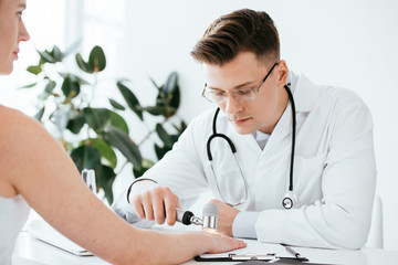 selective focus of dermatologist in glasses examining woman while holding dermatoscope in clinic