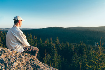 Young man hiker in white cap and shirt sit on rock from back looking to valley