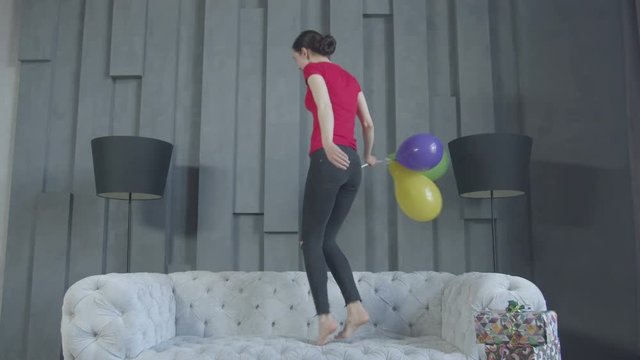 Joyful adult slim woman jumping on couch with colorful balloons in hand enjoying celebration of 30th anniversary at home. Excited beautiful female having fun after birthday party in domestic room.