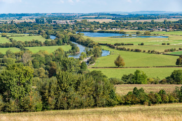 Fototapeta na wymiar A sunlit rural scene with green fields and a river, seen from above. It is the River Thames seen from the hills known as the Wittenham Clumps, in Oxfordshire, United Kingdom. 