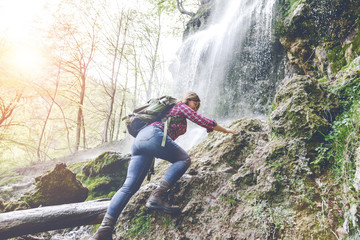 young woman with backpack hiking near waterfall in forest 