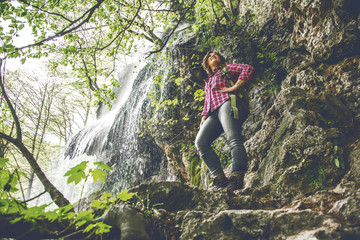 young woman with backpack hiking near waterfall in forest 