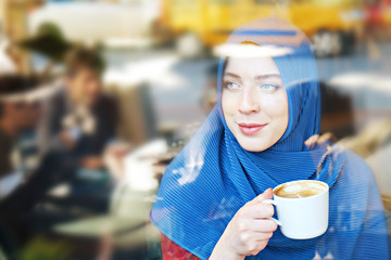 Young beautiful caucasian woman wearing traditional muslim headscarf in hipster coffee shop with big full length windows. Female in blue hijab at cozy cafe. Background, copy space, close up portrait.