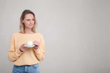 Beautiful young tender girl with blond hair standing with a cup of coffee in her hands, she is dressed in a yellow sweater. poses against a gray background. facial expression, emotion.
