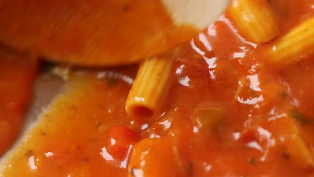 Very close video of rigatoni pasta and slices of sausage in a tomato sauce being stirred with a wood spoon in a pan while heating illuminated with natural lighting.
