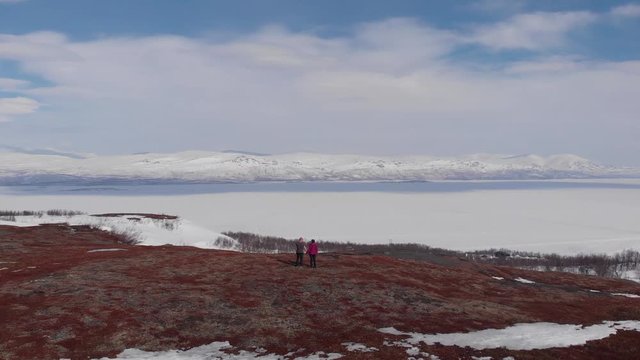 Aerial footage of two people holding hands on top of a mountain in Swedish Lapland with snowy mountains and frozen lake in the background