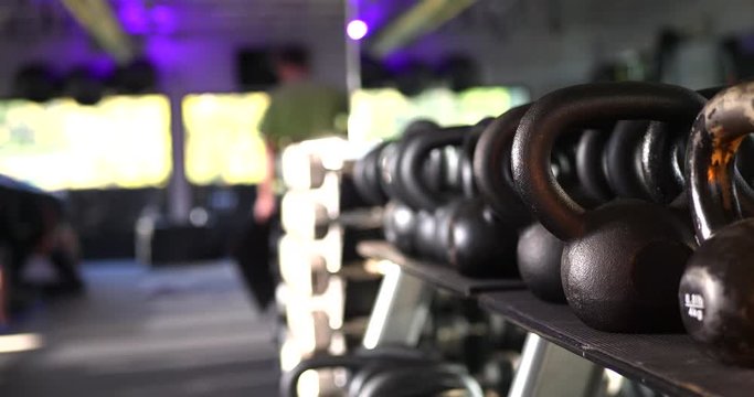 A rack of weights and kettlebells in a gym with people defocused working out and training in the background.