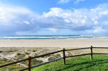 Fototapeta premium Beach with grass, wooden fence and industrial port. Sea with waves and cloudy sky, Sabon, Spain.